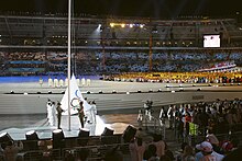 Au premie plan, le drapeau olympique en phase d'être hissé sur un mat. Dans le fond, ds athlètes devant la tribune opposée.