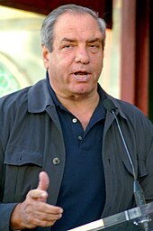 An older man wearing a light jacket over a blue shirt. He is looking toward the camera while speaking in a microphone.