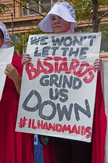 A woman in a long red dress with a white bonnet holding a protest sign