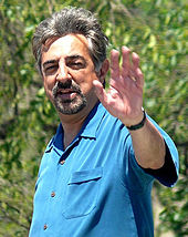 Colored photograph of a man, waving with his left arm. He is wearing a blue shirt.
