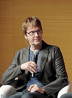 A middle-aged American man holding a microphone wearing glasses, a blue t-shirt, and a black coat