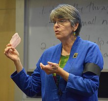 Mary Beth Tinker speaking, holding up a detention slip and wearing a black armband.