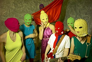 7 women with bright colored clothes and multicolored knit ski masks over their faces. A woman at the center holds a guitar and one at the back holds a piece of red fabric.