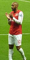 Thierry Henry, wearing a red shirt with white long sleeves and shorts with a number 12 and Nike logo on the left-leg side, applauds.