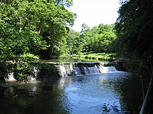 a river flowing over a weir, trees surrounding on a sunny day, blue sky, dark pool in foreground