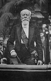 Photo en noir et blanc d’un homme chauve, à la barbe blanche, en tenue officielle, un chapeau dans la main droite