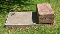 A block of orange-pink stone on a concrete bed, surrounded by grass, with the name "Stallard" carved into the side facing the viewer