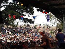 Photo is shot from behind the band looking out to the audience. Two band members are in the foreground at the right. First man (Mikey) is partly turned to his left while playing his guitar. Second man (Huntley) is shown from behind. The other band members are out of view.