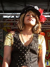 M.I.A. standing in a golden t-shirt, coat and a hat rimmed with flowers.
