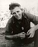 A smiling young man in military uniform, crouching with his forearms resting on his legs. His soft cap is pushed high up on his forehead and his dog tags hang out of his short-sleeved button shirt.