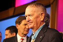 Head and shoulders of an older man with thinning hair wearing a dark suit coat, white shirt, and red tie. A star-shaped medal hangs from a light blue ribbon around his neck and on his lapel is a small round blue pin.