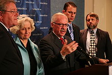 Barr gestures his right arm as his left hand rests on a podium, with two people on each side