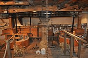 Interior view of the machinery on the first floor. The "Great Spur Wheel" is an iron gear-wheel arranged horizontally on a vertical spindle, which spindle passes through the floor and is driven from below by the mill. on either side are millstones, set in a wooden drum-shaped housing. They are driven by vertical iron shafts which, at the top, have a small gear mating with the great spur wheel. The drive is thereby geared up to a much higher speed than the main vertical shaft from the wheel The floor is bare planks, the walls and apparatus are a golden coloured pale wood, and there is ancient whitewash on the walls. The metal parts of the mechanism are painted a gloss black.