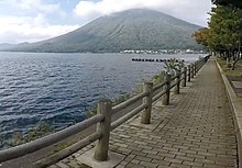 Photo couleur d'un chemin pavé (à droite), le long de la rive d'une étendue d'eau. L'arrière-plan est constitué d'une montagne boisée, sous un ciel bleu nuageux.