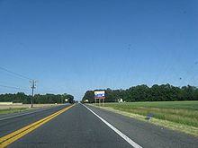 A two-lane road passing through farm fields. A blue and white sign on the right indicates an ongoing construction project on US 113.