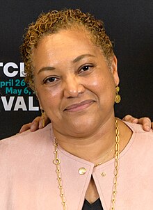 This image shows a smiling woman in her fifties with light brown hair, a golden necklace and a pink suit. She is standing in front of a black background with white and turquoise writing on it.