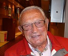 A colour photograph of a Scandinavian man in his eighties. He is seen wearing a red overcoat.