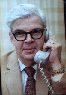 A white-haired man, wearing horn-rimmed glasses and a tan suit, holds a telephone in his left hand.