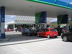 Cars queuing up at a filling station. A typical motorway rest area café and convenience store are visible in the background.