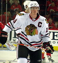 Hockey player in white uniform. He holds looks forward and to his left, holding his stick at waist level.