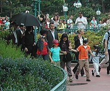 A man in public is dressed in black. He is surrounded by an entourage and members of the public, some of whom are holding cameras. He walks behind three young children, all of whom are wearing facial masks.