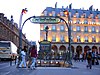 Photographie en couleur et en plein jour d'une bouche de métro dont les candélabres diffusent leur lumière rouge, de même qu'est illuminée la façade de l'hôtel derrière tandis que des voitures circulent à gauche de l'image