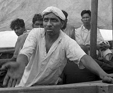 alt = A black-and-white photograph of an Indian sailor aboard a lifeboat, pleading for water.