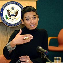 Salbi speaking into a microphone, the United States Department of State crest is in the background. She is dressed in black, with short hair, looking confident and making hand gestures.