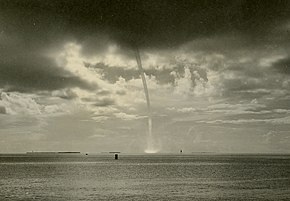 A sepia toned image of a waterspout. The waterspout extends from the dark cloud base at the top of the image to the sea below. Some spray is visible where the waterspout contacts the sea surface, and in the background, partly sunny skies are visible.