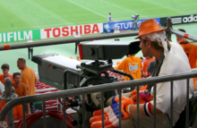 Photo de profil d'un cameraman en tribune filmant le terrain de football.