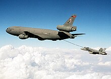 A jet aircraft refuels from a gray three-engine tanker via a long boom located under the tanker's aft fuselage.