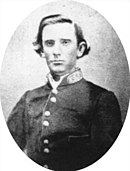 Black and white photo shows a clean-shaven youthful-looking man. He wears a dark gray coat with a single row of buttons and a Confederate general's insignia on the collar.