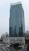 Ground-level view of a blue, glass high-rise; the tower sits behind a small, white, stone, window-dotted facade