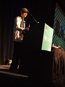 A 25 year old Swedish man in a grey jacket, black pants, and green hat speaking at a conference.