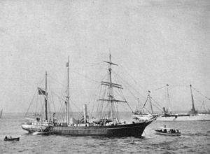 A three-masted ship with sails furled, short funnel amidships, flag flying from the stern on left of picture. Two small boats are close by, and a larger vessel decked with bunting is visible in the background.