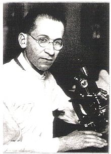 A serious-looking man in circular glasses poses with a microscope.