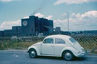 A white Volkswagen Käfer that had just came out of the facility in 1960. Pictured in the background is the factory it was built at.