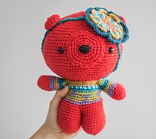 A red stuffed bear, wearing a blue and green sweater, with a flower decoration on the head
