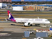 A white twin-engine plane painted with the word "HAWAIIAN" in the front, a gray lei across its fuselage, and a woman in different purple hues on the tail is being towed on an airport taxiway