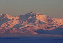 Imposante montagne enneigée, dominant la mer et baignée par une lumière rosée rasante.