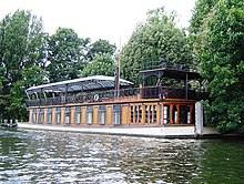 A large, low-slung boat lies moored on the bank of a body of water. The lower deck is made from wood, interrupted by several large windows. The upper deck is made from decorative metal, and covered with a large glass awning.