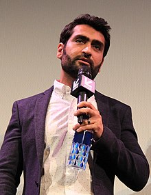 A man dressed in a white shirt and dark jacket speaks into his microphone