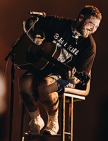 Malone sits on a high chair with a mic and acoustic guitar. A light illuminates his smiling face and casts shadows as he leans camera right.