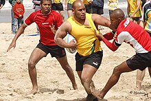 Joueur en jaune, avec le ballon, essayant d'échapper à un joueur qui le tient par le maillot. Un deuxième adversaire est présent sur l'autre côté.