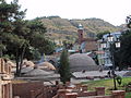 Au pied d'une colline aride, des constructions en briques rouges entourent quelques petits bâtiments en forme de dômes, tandis qu'à l'arrière-plan figure un clocher d'église.