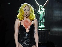 Lady Gaga standing in a black leotard with fake blood on her neck. Behind her a dimly lit fountain statue can be seen