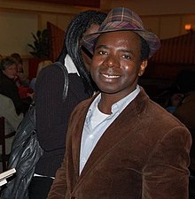 Vamba Sherif wearing a brown jacket over a light shirt, with a plaid-patterned fedora hat, smiling at camera