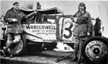 A white man and woman standing in front of a Ford automobile painted with the words "Wanderwell around the world endurance contest"; both are wearing uniform-style clothing