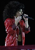 Lady Gaga in a red leather jacket and bushy black hair singing.