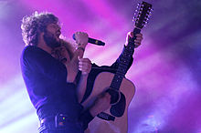Upper body shot of two men performing. Littlemore is on the left and is shown in right profile, he is holding a microphone in his right hand and singing with his eyes closed. He wears a dark blue shirt, dark pants. At his right hip in his belt is an electronic device. The second man is partly obscured, he leans in close to the first. He is singing and playing a 12-string guitar with his left hand high on the fret board. His right hand holds the guitar pick as he strums the strings. His eyes are open and looking at Littlemore.